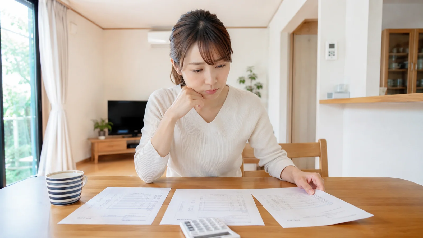 リビングのテーブルで複数の見積書類と電卓を見ながら検討する女性。料金比較や見積もり診断のシーンに最適な落ち着いた室内写真。